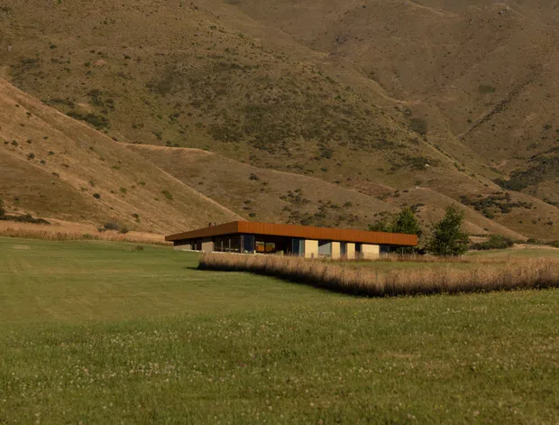 A low-profile modern building set within an expansive rural meadow, backed by dry rolling mountain slopes. The structure has a long horizontal form, dark glazing, pale walls, and a distinctive warm rust-orange roof or fascia, with a sparse landscaped edge.