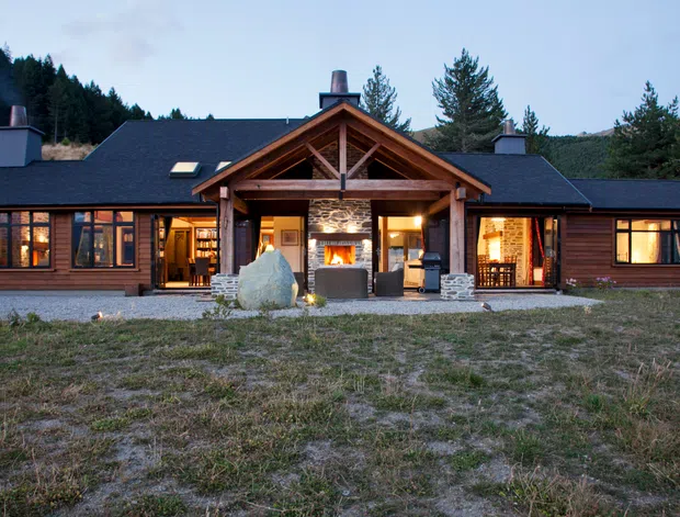 Single-storey lodge-style house set against a forested hillside, with dark timber cladding, a broad gabled roof, multiple chimneys, and a prominent covered timber-framed entry or outdoor living area. Warm interior lighting is visible through the windows and open central doors, with a stone fireplace and large natural boulders at the entrance.