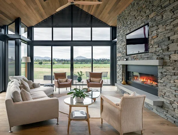 A spacious, light-filled living room with a dramatic vaulted timber-lined ceiling, exposed gable form, black-framed floor-to-ceiling glazing, and a rugged stacked-stone fireplace wall. Neutral upholstered sofas and armchairs surround round coffee tables, while a large ceiling fan hangs centrally. The glazing opens views toward fields and distant hills, creating a warm, refined indoor-outdoor atmosphere.