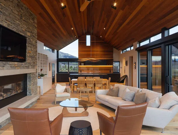 A spacious, open-plan living room with a dramatic pitched timber-lined ceiling, extensive dark-framed glazing, a large stacked-stone fireplace wall, and an integrated kitchen and dining area. The room is furnished with a pale sectional sofa, leather lounge chairs, a glass coffee table, and timber dining furniture. The glazing opens toward a wooded or mountainous landscape, while warm timber and stone finishes create a refined lodge-like atmosphere.