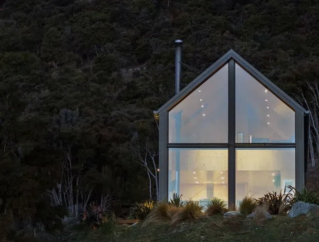 A compact contemporary cabin-like house set into a heavily wooded hillside, photographed at dusk. The front elevation is dominated by a steep gabled form with extensive floor-to-roof glazing divided by dark structural framing. Warm interior lighting is visible through the windows, with a chimney rising beside the roof and low native-style planting around the base.
