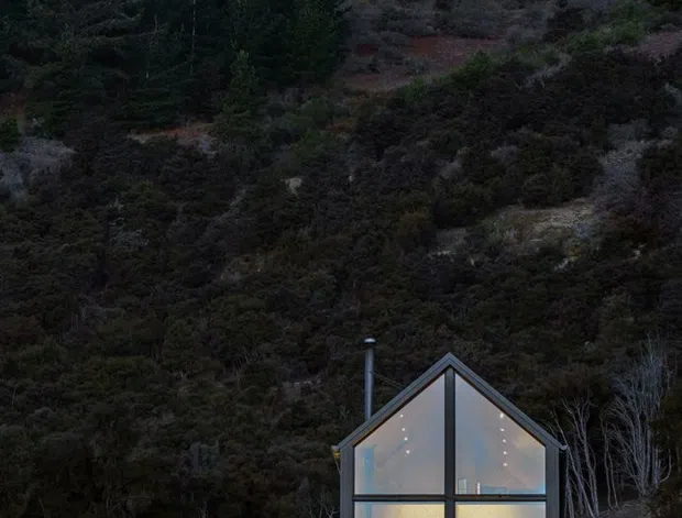 A small, steeply gabled glass-fronted dwelling sits against a densely wooded hillside at dusk. Dark structural framing divides the full-height glazing, with warm interior lights visible through the facade. The building is nestled into a rugged, sloped natural site surrounded by trees and scrub.