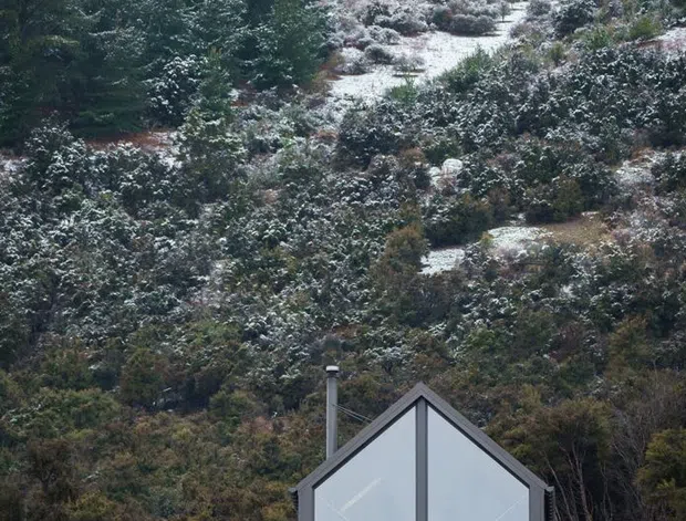 A small contemporary gable-front cabin or house set against a steep, densely vegetated hillside with light snow cover. The compact facade is dominated by large grey-framed glazed panels, with a simple grey roof edge and a visible metal flue. Native grasses and low planting surround the building, creating a secluded alpine or mountainous setting.