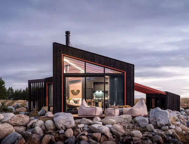 A compact modern house set among a rugged field of large rocks, viewed from outside at dusk. The dark, rectilinear facade features extensive floor-to-ceiling glazing, a dramatic sloping roofline, and warm interior lighting. Outdoor lounge seating is arranged in front of the glazed living area, with a sparse natural landscape visible around the building.