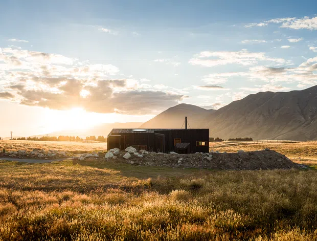A small dark-clad single-storey cabin or dwelling set in an open alpine or rural landscape, with rugged mountains rising behind it. The low, simple building has a shallow pitched roof, dark vertical cladding, and a rocky perimeter. Warm low-angle sunlight creates a golden, dramatic atmosphere.