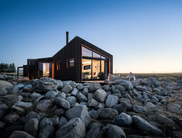 A compact dark-clad modern cabin set among a broad field of rounded rocks. The single-storey building has a pitched roofline, large glazed openings with warm interior light, and a small outdoor seating area beside the main window. The sparse rocky setting and strong contrast between the dark exterior and illuminated interior create a remote, atmospheric scene.