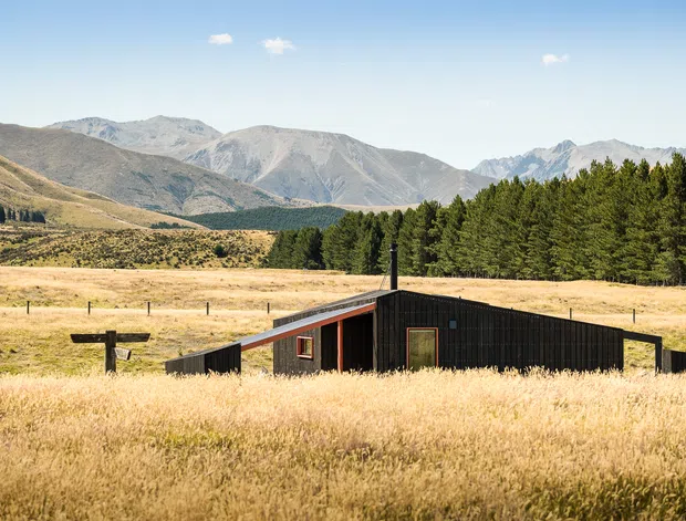 A low-profile, dark-clad architectural building sits within a golden grassland, with a strong angular roofline and simple rectangular openings. A dense stand of trees and expansive rugged mountains form the background under a clear blue sky, giving the scene a secluded alpine rural character.