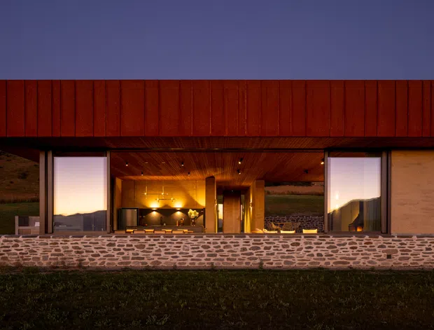 A low, modern building viewed from outside at dusk, set against an open rural hillside. The façade has a long horizontal form with deep red-brown vertical cladding above a broad opening, large glazed panels, and a rough stone masonry base. Warm timber-lined ceilings and interior lighting are visible through the open frontage, creating a strong indoor-outdoor connection.