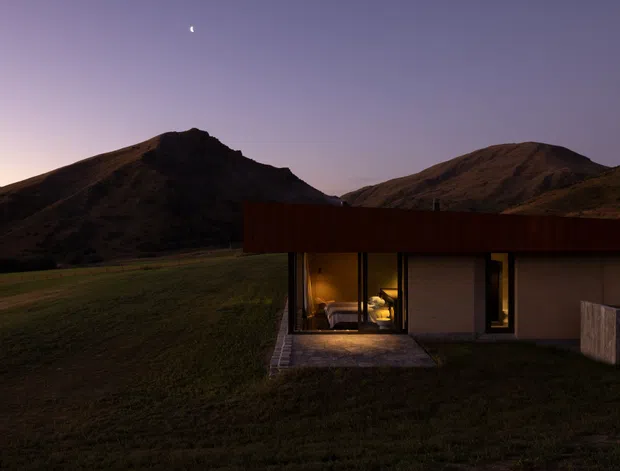 A low, modern single-storey dwelling sits in a broad grassy alpine or mountainous landscape at dusk. The building has a simple flat roof with a dark rust-toned fascia or cladding, pale rendered walls, large warm-lit windows, and a small paved entry terrace. Steep, rugged hills rise directly behind the house beneath a dusky purple sky with a crescent moon.