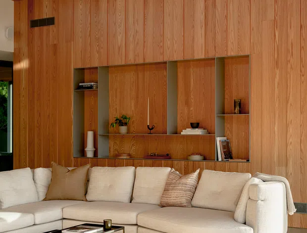 A warm, refined living room featuring a large cream sectional sofa, nested metal coffee tables, and a full-height built-in timber wall with integrated shelving and concealed storage. The wall and ceiling are clad in richly coloured vertical-grain wood, creating a cohesive architectural feature. Sunlight enters from the side, casting long shadows across the pale rug and floor.