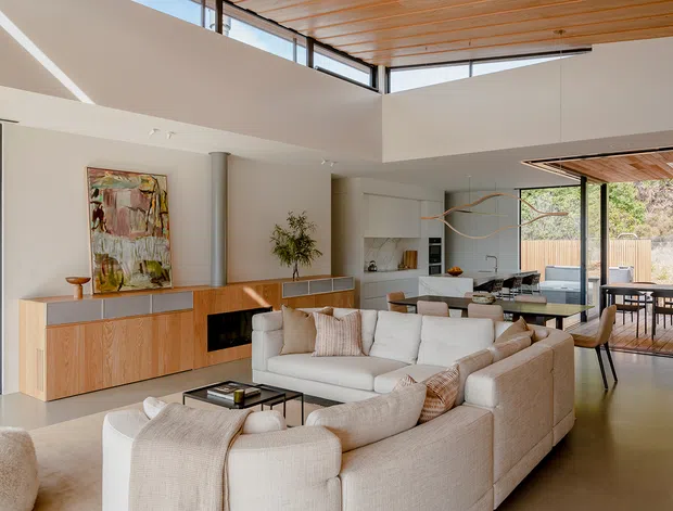 A bright, spacious open-plan living area with a large cream sectional sofa, low black coffee table, and timber media/storage console. The living room opens directly to a modern white kitchen and dining area, with full-height glazing leading to an outdoor deck. Architectural features include a high ceiling, clerestory glazing, warm timber ceiling lining, polished concrete flooring, and restrained contemporary joinery.