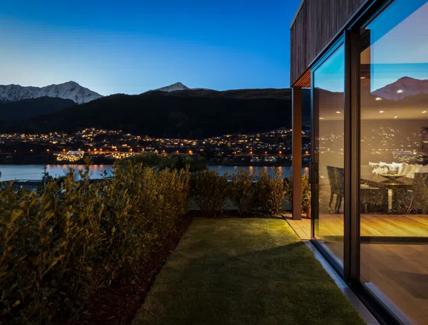 Twilight exterior view beside a contemporary glazed building, looking across a landscaped garden toward a lake and illuminated settlement beneath dark mountain ridges. The building has extensive floor-to-ceiling glazing, dark vertical timber cladding, and warm timber flooring visible inside. Reflections in the glazing show the mountainous landscape and a dining area.