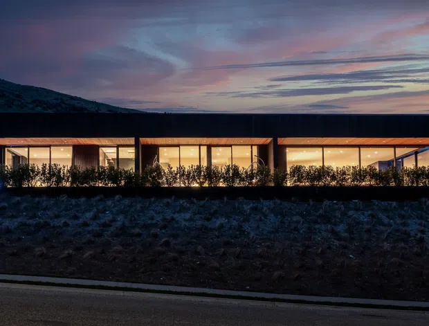 Night-time exterior view of a long, low contemporary building on a sloping, sparsely vegetated site. Three connected or closely spaced glazed sections glow warmly from within, with dark cladding, projecting timber-toned roof edges, and dense low planting screening the facade. A mountain or hillside rises behind the building beneath a dramatic twilight sky, while a road runs across the foreground.