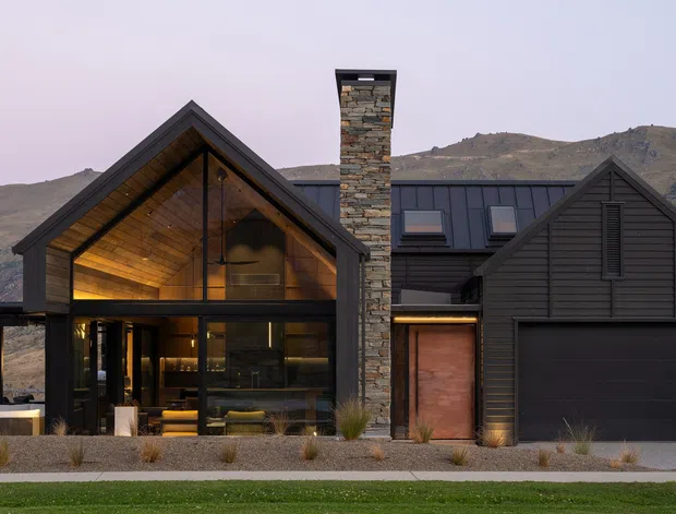 Front exterior view of a contemporary alpine-style residence with dark horizontal cladding, steep gabled forms, extensive floor-to-ceiling glazing, a prominent natural-stone chimney, and an integrated garage. Warm interior lighting is visible through the glass, with a dry landscaped forecourt and rugged mountain terrain behind.
