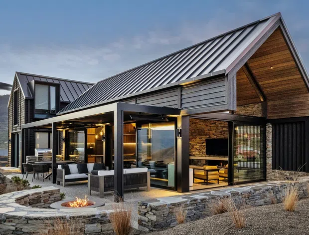 Exterior view of a modern alpine residence with steep pitched metal roofs, dark vertical cladding, timber-lined gable soffits, extensive glazing, and a covered outdoor living area. A landscaped stone terrace includes built-in seating and a small fire pit, with rugged mountainous terrain in the background.