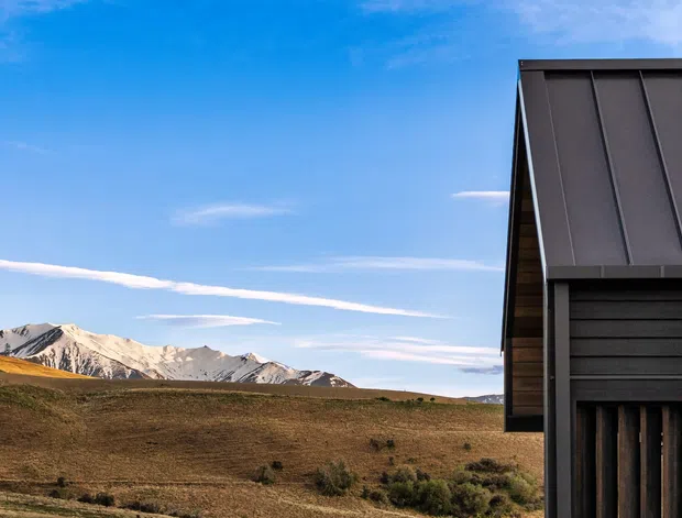 Exterior architectural detail of a contemporary mountain building, showing a dark standing-seam metal roof and cladding beside a rugged stacked-stone wall. The structure is set against an expansive alpine landscape with snow-dusted mountain peaks and open brown terrain.