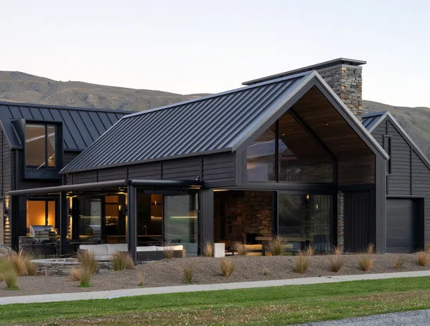 Exterior view of a contemporary multi-level mountain home with intersecting steep gabled roof forms, dark vertical cladding, standing-seam metal roofing, extensive glazing, and a prominent stone chimney. The house is set against a rugged alpine or mountainous landscape, with a gravelled, drought-tolerant landscaped frontage and a garage integrated into the right side.