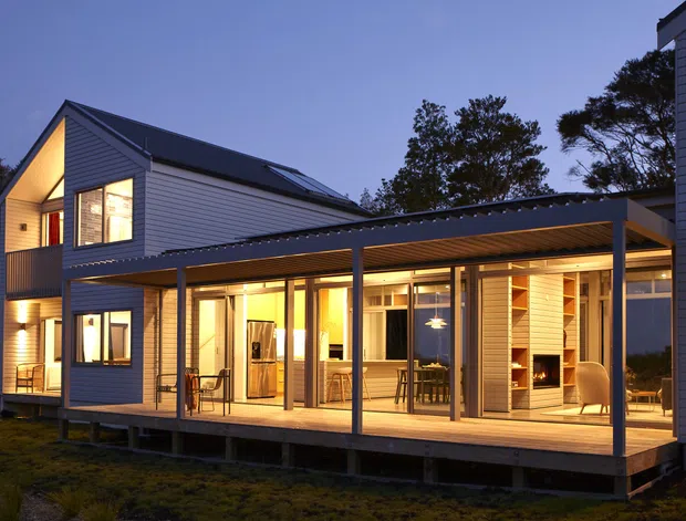 Twilight exterior view of a contemporary multi-level home with grey horizontal weatherboard cladding, dark pitched roofs, extensive glazed openings, and a broad covered timber deck. Warm interior lighting reveals open-plan living and dining areas through the glazing, while the elevated deck and covered outdoor connection are prominent.
