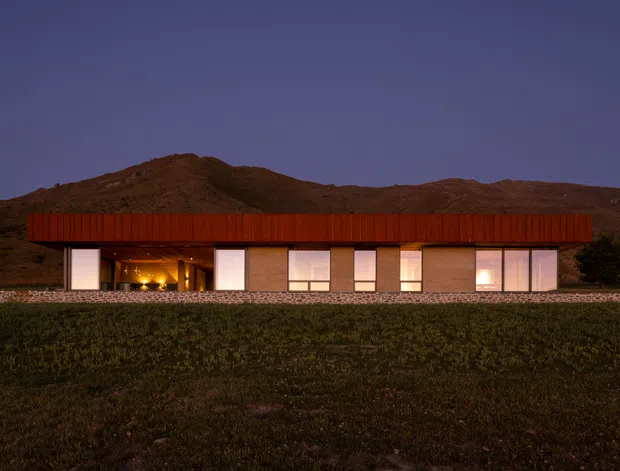 A low, single-storey modern house photographed at dusk, set against a rugged mountainous landscape. The building has a long horizontal form, a broad flat or very low-pitched roof with dark timber-toned cladding, extensive rectangular glazing, warm interior lighting, and a low stone-clad base.