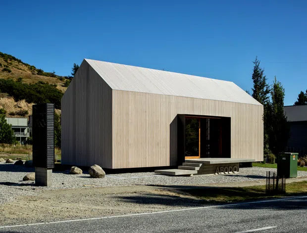 A compact contemporary building with a simple gable-roofed form, clad in vertically ribbed light timber or metal-toned panels. A large recessed dark entry opening faces a gravel forecourt, with concrete steps leading inside. The building sits in a dry, sloping landscape with tall trees and neighbouring structures visible around it.
