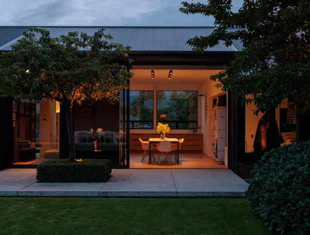 Twilight view of a single-storey contemporary home with a dark corrugated metal roof and a recessed, warmly lit central glazed dining area. The symmetrical facade is framed by dark structural elements and mature landscaping, with a paved terrace or entry apron leading to the dining space. Interior furnishings are visible through the glazing.