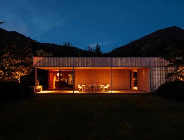 A low, single-storey modern pavilion-style house photographed at dusk, set against dark wooded hills. The building has a long rectangular flat roof, extensive floor-to-ceiling glazing, a warm timber-toned illuminated interior, and a covered outdoor terrace with dining furniture. Vertical slatted screening and timber-like facade elements frame the glazed living area.