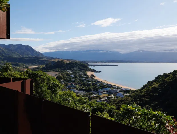 Elevated exterior view from a dark-clad architectural structure overlooking a coastal bay, sandy beach, surrounding suburban settlement, wooded hills, and distant mountains. The image is dominated by the expansive waterfront landscape and framed by dark brown architectural cladding or screening in the foreground.