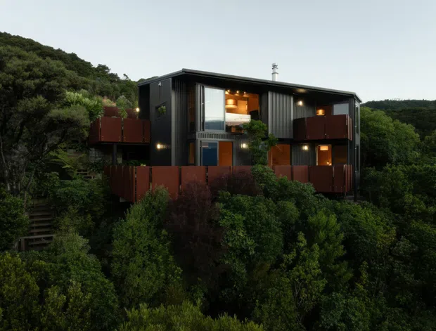 Exterior view of a contemporary multi-level dark-clad house set within dense native vegetation on a steep, elevated site. The building features a low-pitched flat roof, projecting balconies or decks with rust-red privacy screens, dark vertical cladding, large windows, and warm interior lighting visible through the glazing. The surrounding bushland is prominent, and the image is captured in subdued evening light.