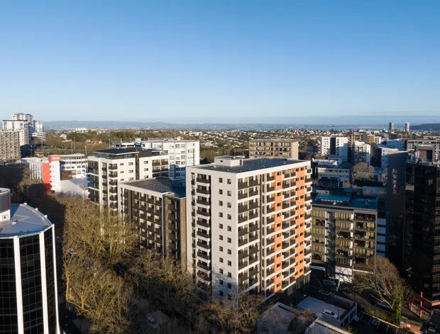 A wide elevated view over a dense urban district, dominated by modern multi-storey apartment and commercial buildings. The foreground includes several mid- and high-rise blocks with flat roofs, balconies, and contrasting dark, white, and orange facades. A broad cityscape extends toward a visible waterfront or large body of water on the horizon under a clear blue sky.