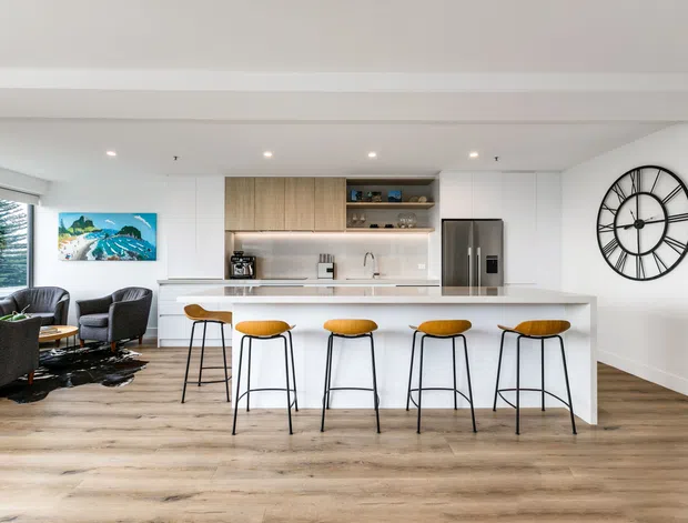Bright open-plan kitchen and living area with a large white island breakfast bar, five timber-and-black bar stools, integrated white cabinetry, warm wood-grain overhead joinery, stainless-steel refrigerator, and pale timber-look flooring. A dark seating area and artwork are visible to the left, with a large black wall clock on the right. Recessed ceiling lighting and broad glazing create an airy contemporary interior.