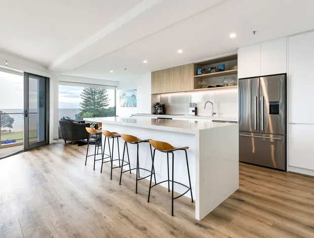 Bright open-plan kitchen and living area with a large white island, four timber-seated bar stools, integrated white cabinetry, stainless steel refrigerator, timber open shelving, and warm timber-look flooring. Full-height glazing and a sliding door open onto a balcony with coastal water views.