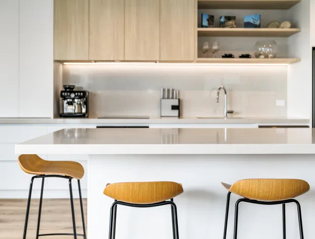 Bright contemporary kitchen featuring pale timber overhead cabinetry, open display shelving, a light stone island benchtop with integrated seating, a pale splashback, stainless-steel tapware and appliances, and three timber-and-black-metal bar stools. The space has a clean, refined and airy appearance.
