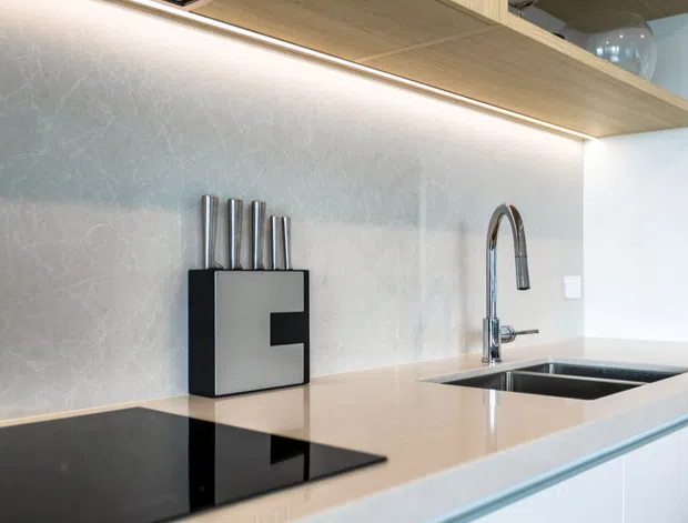 Close-up view of a contemporary kitchen benchtop featuring a pale stone surface, dark glass cooktop, undermount double sink, chrome gooseneck tap, and light timber overhead shelving with integrated under-shelf lighting. A pale textured splashback and white cabinetry create a clean, refined appearance.