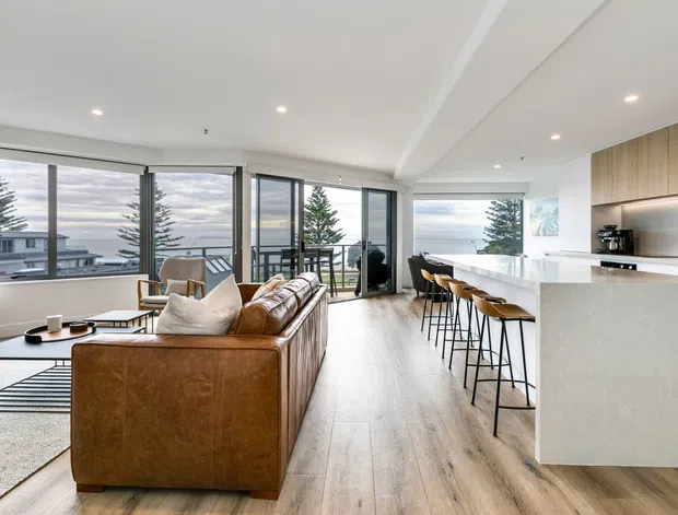 Open-plan contemporary living, dining and kitchen area with floor-to-ceiling glazing, pale timber flooring, a large brown leather sectional sofa, and a white island with bar seating. The room overlooks a coastal or suburban landscape and is finished with light wood cabinetry, recessed lighting and a bright neutral palette.
