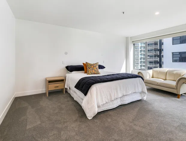 Bright, spacious contemporary bedroom with a large bed, neutral bedding, grey carpet, a simple timber bedside table, and a cream upholstered sofa positioned beside expansive apartment windows overlooking nearby high-rise buildings. White walls and ceiling create a clean, airy interior.