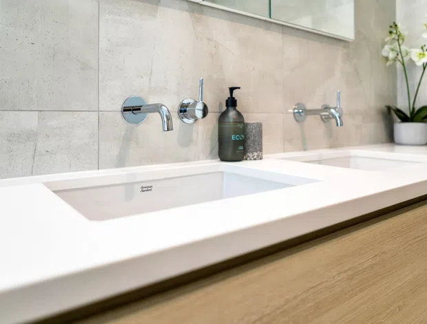 Close-up of a contemporary bathroom vanity with a wide white double basin countertop, wall-mounted chrome tapware, pale large-format tiled walls, and light timber cabinetry. A small orchid and soap dispenser provide subtle styling accents.