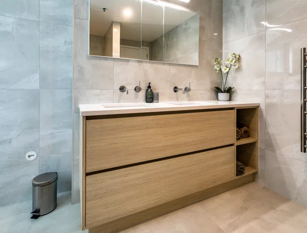 Contemporary bathroom with large pale stone-look wall and floor tiles, a floating timber-look double-drawer vanity, white benchtop basin, wall-mounted taps, mirrored cabinet, stainless-steel heated towel rail, and recessed shelving. The neutral, clean-lined fit-out is softly illuminated and includes a small orchid and folded towels.