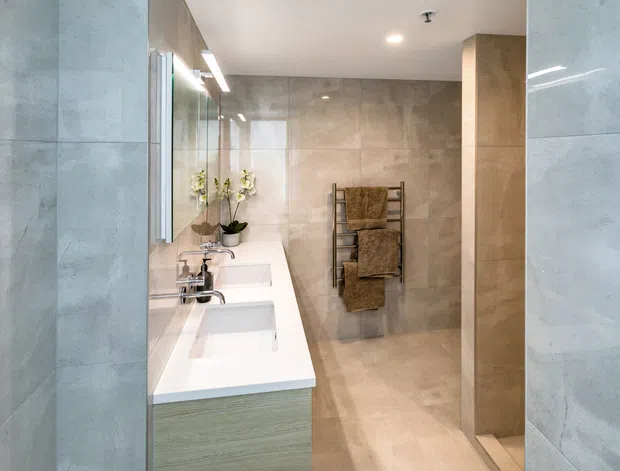 A contemporary bathroom viewed through a tiled opening, featuring a long white double vanity with integrated basins, wall-mounted mirrors, pale timber-toned cabinetry, large beige stone-look tiles, recessed ceiling lighting, and a towel ladder in the shower area. The space has a clean, warm-neutral and highly streamlined appearance.