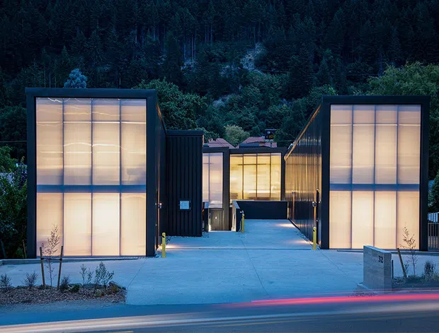 Twilight exterior view of a contemporary multi-building complex set against a dense forested hillside. Several compact, flat-roofed structures line a broad concrete forecourt and central pedestrian approach. The buildings feature dark metal cladding, large translucent illuminated wall panels, and crisp rectilinear forms, with motion-blurred vehicle lights along the street in the foreground.