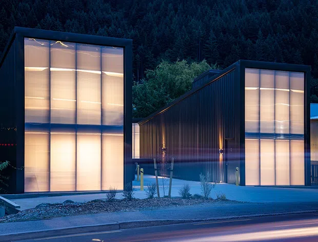 Dusk exterior view of a contemporary multi-volume building in a forested setting. The composition features dark, sharply geometric façades with tall translucent illuminated glazing, a steeply sloping roof form, vertical metal-like cladding, and a paved street frontage. Warm interior light contrasts with the cool blue evening atmosphere and surrounding woodland.