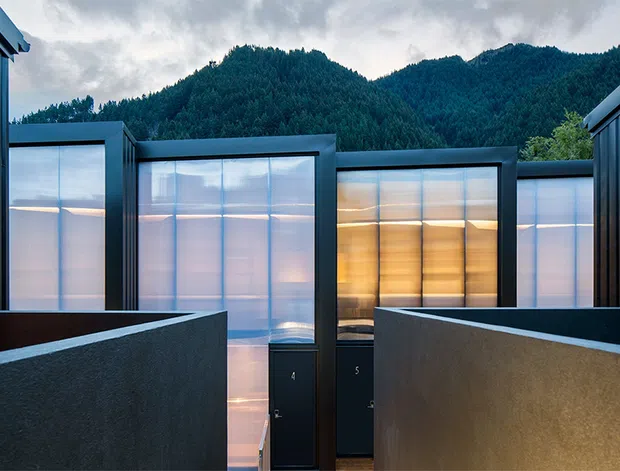 Exterior architectural view of a contemporary multi-volume building set against densely forested mountains. Dark vertical metal cladding and flat-roofed forms frame translucent illuminated wall panels, with tall boundary walls and a central recessed entry creating a strong geometric composition.