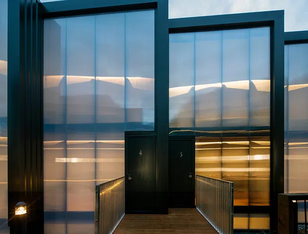 Exterior view of a contemporary multi-unit building with tall translucent glazed facade panels framed in dark metal. A timber walkway with metal balustrades leads to numbered dark entrance doors, with warm interior lighting visible through the glazing.