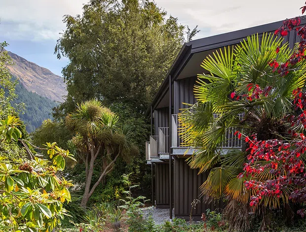 Exterior view of a dark-clad, contemporary multi-level building nestled among dense landscaped planting, with balconies and extensive glazing visible along the side. A steep mountainous backdrop is partially visible beyond the vegetation.