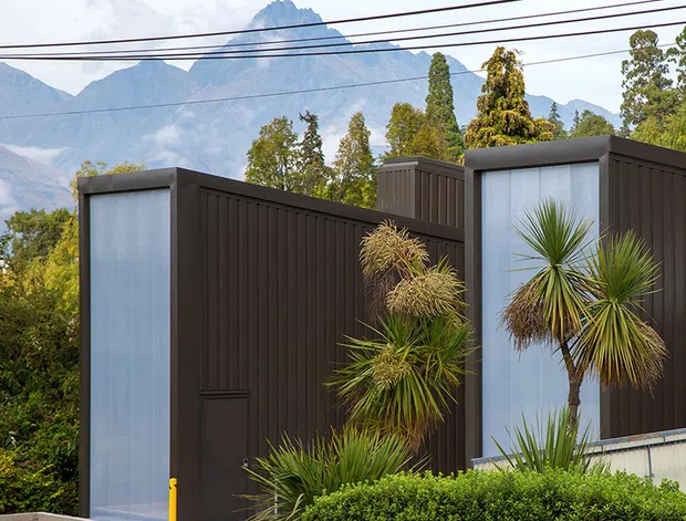 Exterior view of a contemporary dark-clad building with sharply composed rectilinear volumes, tall translucent glazed panels, and spiky architectural planting in the foreground. Dramatic mountain peaks and mature trees form the background, while overhead utility wires cross the upper portion of the image.