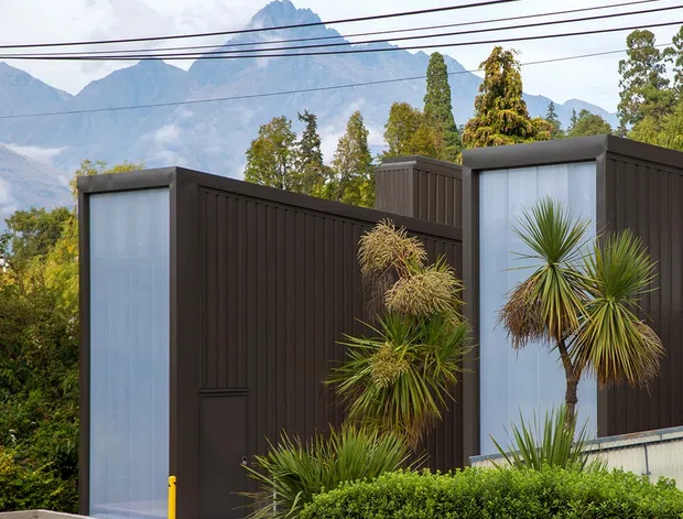 Exterior view of a contemporary dark-clad building with tall translucent glazed panels and sculptural architectural volumes. Layered native and drought-tolerant planting softens the facade, while dramatic mountain peaks and overhead utility wires form the background.