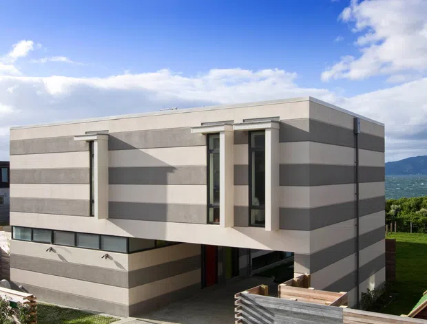 Exterior view of a contemporary two-storey rectangular home with a flat roof and bold horizontal bands of light and dark grey cladding. Narrow vertical windows and projecting architectural fins create strong geometric shadows. A partially enclosed lower level, timber fencing, landscaped lawn, and glimpses of water and mountains are visible in the surrounding setting.
