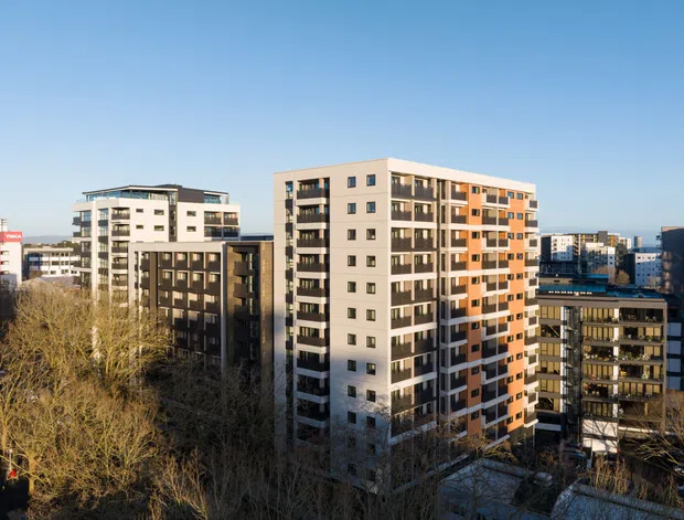 Urban exterior view of several contemporary multi-storey apartment buildings, with prominent mid-rise residential blocks featuring balconies, varied window arrangements, and light neutral façades accented by dark grey and orange panels. Leafless trees occupy the foreground and a clear blue sky frames the city skyline.