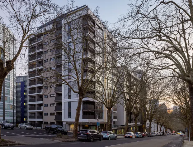 Street-level exterior view of a multi-storey apartment building along a broad, tree-lined urban boulevard. The building has a contemporary facade with balconies, dark and light rendered panels, and extensive glazing, with neighbouring apartment and commercial buildings visible nearby. Parked cars and mature deciduous street trees frame the scene.
