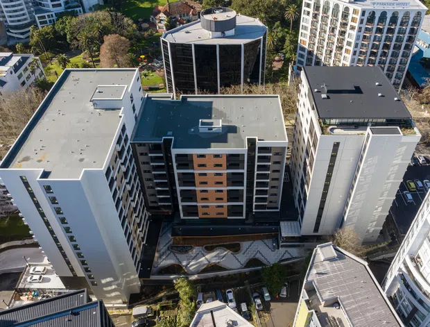 Aerial view of a dense urban multi-storey residential or commercial precinct, showing several contemporary high-rise buildings with flat roofs, rooftop plant and equipment, glazed facades, balconies, landscaped courtyards, and surrounding city development.