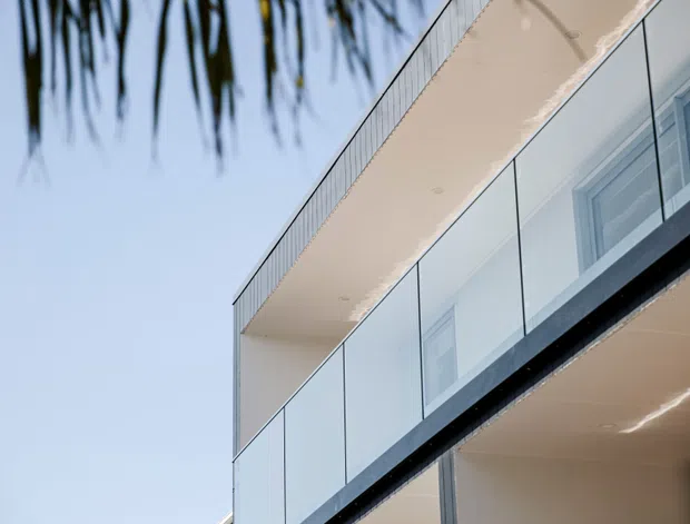 Close-up exterior view of a contemporary multi-level building, featuring broad projecting balconies with clear glass balustrades, pale rendered or panelled surfaces, and large glazed openings. Palm fronds frame the upper left, suggesting a warm coastal or suburban setting. The image focuses on the building facade and balcony architecture.